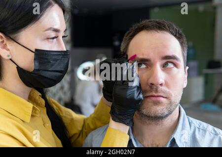 Ein junger Mann über das Verfahren der Augenbrauenkorrektur in einem Schönheitssalon. Die Meisterin entfernt einem Typen eine Wimpern. Männer werden die Augenbrauen zupfen. Stockfoto