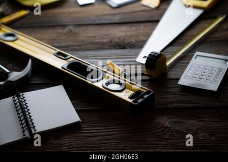 Konzept des Auftragnehmers. Werkzeuge des Auftragnehmers: Gelber Hardhut, libella, Handsäge und Notizbuch auf dem rustikalen Holztisch. Stockfoto