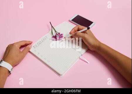 Draufsicht auf weibliche Hände mit Bleistift, Schreiben im Notizbuch, Überprüfung der To-Do-Liste. Handy und rosa Blume liegen auf rosa Hintergrund mit Kopie Spa Stockfoto