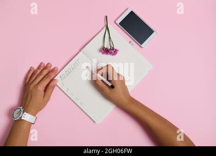 Flaches Blatt weiblicher Hände mit Bleistift, Schreiben im Notizbuch, Überprüfung der To-Do-Liste. Handy und rosa Blume liegen auf rosa Hintergrund mit Kopie Spa Stockfoto