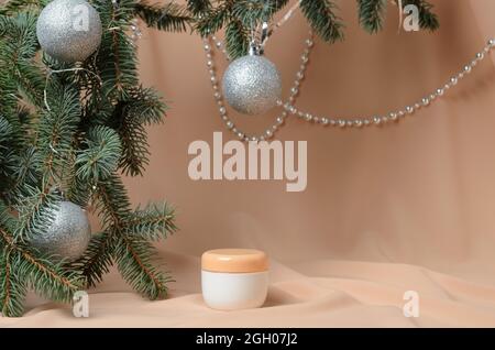 Neujahrsdekoration. Fichtenzweige mit silbernen Kugeln, Hautcreme in einem weißen Glas auf einem beige welligen Hintergrund aus Chiffon. Selektiver Fokus. Stockfoto