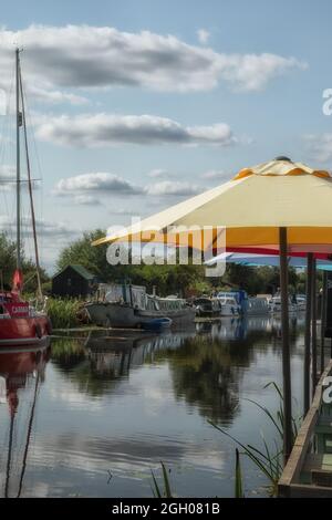 EYBRIDGE BASIN, ESSEX, Großbritannien - 25. AUGUST 2021: Blick auf festgetäute Boote auf der Chelmer und Blackwater Navigation unter Regenschirmen in einem Riverside Cafe in s Stockfoto