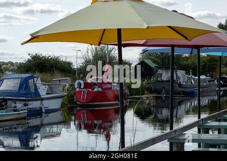 HEYBRIDGE BASIN, ESSEX, UK - 25. AUGUST 2021: Im Sommer werden Boote auf der Chelmer und Blackwater Navigation unter Regenschirmen im Riverside Cafe vertäut Stockfoto