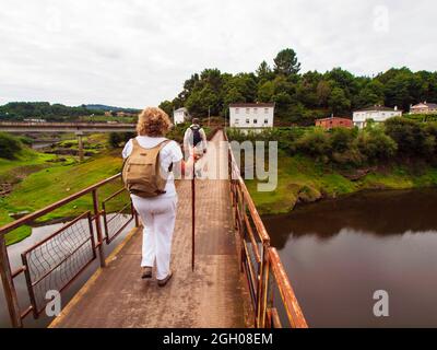 Pilgerin, die über eine steinerne Brücke mit zwei gelben Pfeilen zur Orientierung auf der Route des Camino de Santiago durch Galizien fährt. Stockfoto