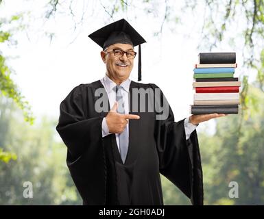 Reifer Mann in einem Abschlusskleid mit einem Stapel von Büchern und zeigt, posiert in einem Park im Freien Stockfoto