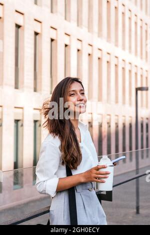 Vertikales Porträt einer attraktiven jungen inderin. Sie steht in der Nähe von Gerichtsgebäuden und hält eine Thermoskanne mit Kaffee und Telefon Stockfoto