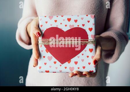 Nahaufnahme einer Geschenkbox, die mit einem roten Herz in den Händen einer nicht erkennbaren Frau verziert ist. Stockfoto