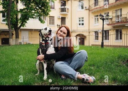 Glückliche Frau posiert und spielt mit ihrem dalmatinischen Hund, während sie im grünen Gras während eines Stadtspaziergangs sitzt. Freundschaft, Liebe und Fürsorge Konzept Stockfoto