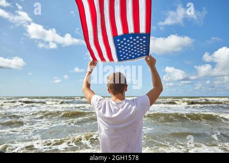 Mann in weißem Outfit, der mit einer amerikanischen Flagge am Strand im Wind winkt. Stockfoto