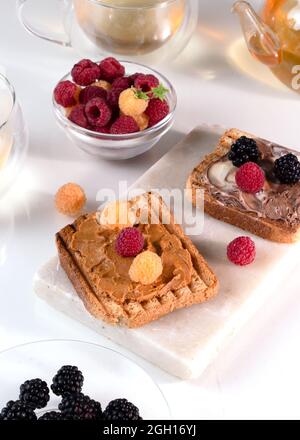 Gesundes Frühstück. Sandwiches oder Toast mit Erdnussbutter, Schokoladenpaste und gelben und roten Himbeeren, Brombeere auf weißem Tisch. Vertikale Aufnahme. Stockfoto