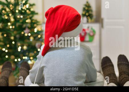 Familie zu Weihnachten am geschmückten Weihnachtsbaum. Ein Kind im Weihnachtsmann-Hut zwischen seinen Eltern in Socken mit Ornamenten. Stockfoto