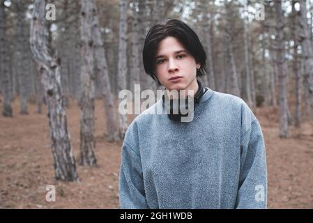 Teenager, 16-17 Jahre alt, tragen Sweatshirt und Kopfhörer, posieren im Wald und schauen auf die Kamera. Trauriger, ernsthafter Kerl im Freien. Stockfoto