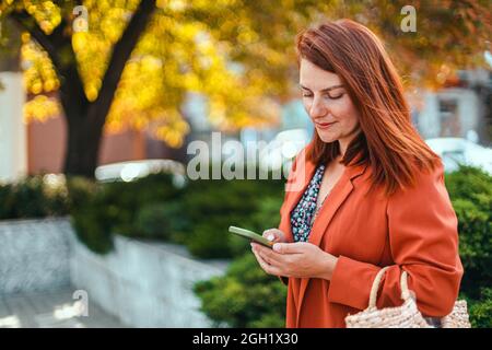 Schöne Geschäftsfrau in stilvoller Kleidung in den Händen nutzt Smartphone-Gerät in einem Stadtpark in einem schönen sonnigen Herbstwetter Stockfoto