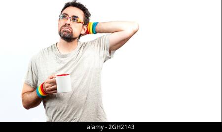 Dunkelhaariger Mann in grauem Hemd mit Brille und einem Armband in Regenbogenfarben; mit müdem Ausdruck, mit einer Hand, die eine Tasse hält. LGBTQ-Konzept. Stockfoto