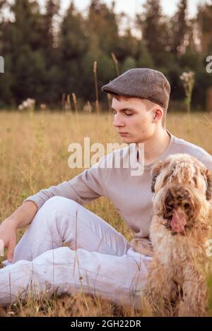 Ein 18-jähriger Junge mit Mütze sitzt an einem Sommerabend mit einem Hund auf einem Feld im dicken Gras. Stockfoto