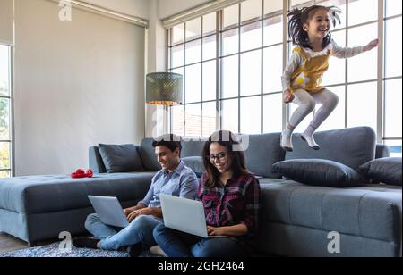 Mischen Sie Rennfamilienliebhaber, kaukasischen Ehemann und asiatische Frau, Eltern arbeiten mit Laptop-Computern auf dem Boden, während kleine freche Mädchen spielen und springen so Stockfoto