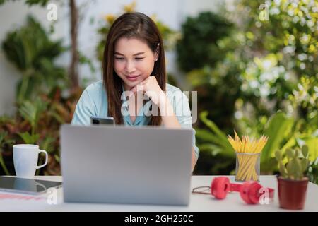 Geschäftsfrau sitzt am Schreibtisch mit Smartphone und Laptop-Computer verbinden sich mit dem Internet in kleinen Garten grünen Hintergrund Ecke zu Hause. Concep Stockfoto