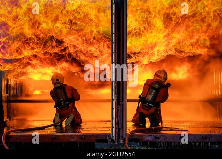 Team von zwei Feuerwehrleuten tragen volle Uniform mit Sauerstoff-Tank auf der Rückseite halten Rohr verteilt Wasser und Gesicht zu breiten riesigen orange heißen Feuer mit tapferen und Stockfoto