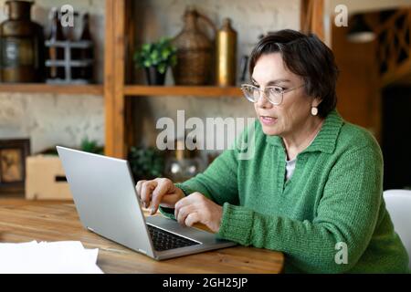 Edly Woman arbeitet zu Hause vor einem Laptop-Monitor mit Papieren. Heimbüro. Sie geht durch Steuererklärungen und prüft Rechnungen. Stockfoto