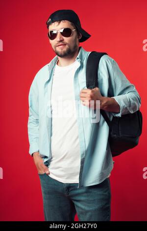 Ein europäischer Tourist in einer Baseballmütze mit Rucksack auf der Schulter steht auf rotem Hintergrund. Ein bärtiger Reisender in legerer Kleidung. Stockfoto