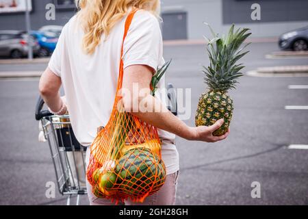 Frau mit Netzbeutel, die Ananas auf einem Supermarkt-Parkplatz hält. Weibliche Person mit Einkaufswagen nach dem Kauf von Lebensmitteln. Keine Verschwendung und Nachhaltigkeit Stockfoto