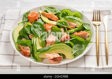 Lachs Spinat Avocado und grüner Pfeffer Salat auf einem weißen Teller mit goldener Gabel und Messer auf einem weißen Holztisch, mediterrane gesunde Ernährung, horizonta Stockfoto