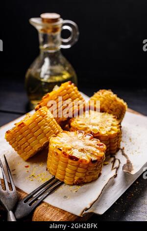 Nahaufnahme gegrillter Mais auf dem Maiskolben mit Öl und Salz auf einem Holzbrett, Nahaufnahme auf dunklem Holzhintergrund. Köstlicher gegrillter Mais und eine Flasche Stockfoto