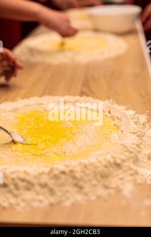 Die Hände mehrerer Personen kneten den Teig auf dem Tisch. Ein rohes Ei mit Mehl vermischen. Nahaufnahme. Stockfoto