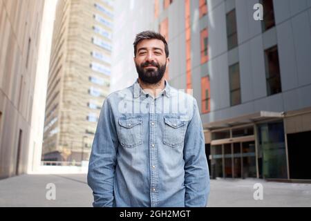 Attraktiver Geschäftsmann, der die Kamera im Freien in der Nähe des großen Bürogebäudes ansieht. Junge lässig gekleidete junge Mann mit fröhlichem Lächeln, Unternehmer Stockfoto