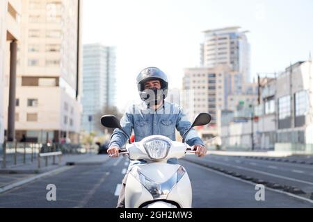 Die mittlere Aufnahme eines jungen Motorradfahrers hielt an einer Ampel in Barcelona an. Der Mann fährt mit seinem weißen Roller auf einer großen Allee voll durch die Stadt Stockfoto