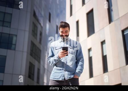 Junger Geschäftsmann im Denim-Hemd mit Smartphone in der Nähe von Bürogebäuden im Finanzzentrum der Stadt. Stockfoto