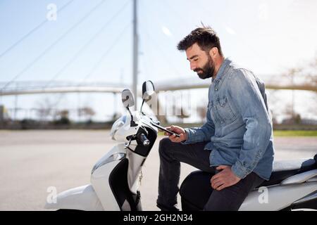 Junger Spanier, der Handy oder Smartphone benutzt und auf seinem Motorroller draußen in der Stadt sitzt. Geschäftsmann überprüft Arbeitsprobleme auf dem Handy. Stockfoto