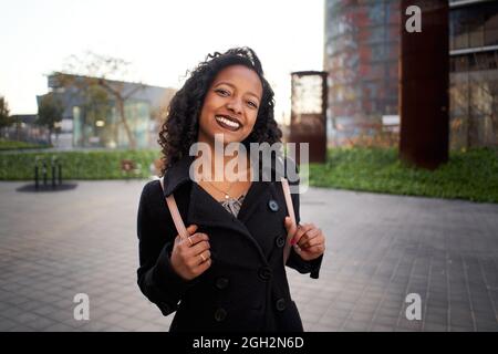 Die Geschäftsfrau von Latina lächelt fröhlich und schaut auf die Kamera. Die junge Frau steht neben einem modernen Bürogebäude in der Stadt. Sie trägt einen Stockfoto