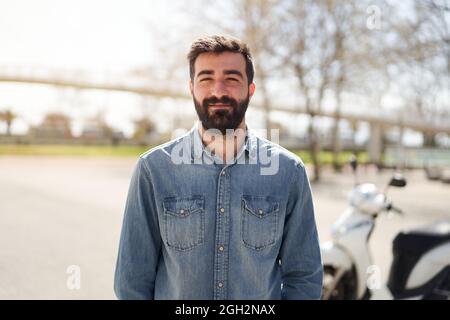 Porträt eines jungen Spanierers, der die Kamera anschaut. Der Mann mit fröhlichem Lächeln steht vor seinem Motorrad, das in der Stadt abgestellt wird. Stockfoto