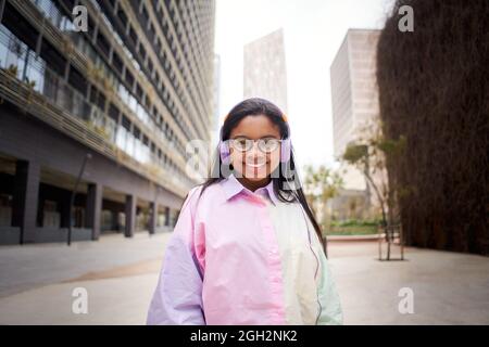 Junge, lächelnde Afroamerikanerin, die farbenfroh gekleidet in die Kamera schaut und Musik mit Kopfhörern auf urbanem Hintergrund in der Stadt hört. Stockfoto
