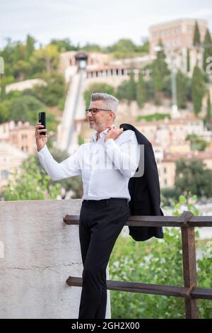 Geschäftsmann, der Videokonferenzanruf auf dem Smartphone macht und auf der Straße redet und auf den Bildschirm schaut. Glücklicher Mann, der Selfie mit dem Handy im Freien macht Stockfoto