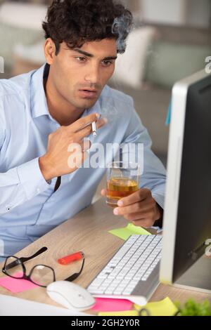 Geschäftsmann an seinem Schreibtisch, der Alkohol trinkt und Zigarette raucht Stockfoto