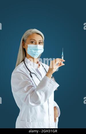 Silberhaarige asiatische Therapeutin mit Maske hält Spritze mit Medizin auf blauem Hintergrund Stockfoto