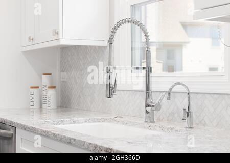 Ein Waschbecken, das in einer renovierten Küche mit weißen Schränken, Chromwasserhahn, Marmor-Fischgrätenmuster-Backplash und Granitarbeitsflächen fotografiert wurde. Stockfoto