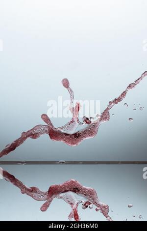 Vertikale Aufnahme von Wein, der auf einer reflektierenden Glasoberfläche vor grauem Hintergrund spritzt Stockfoto