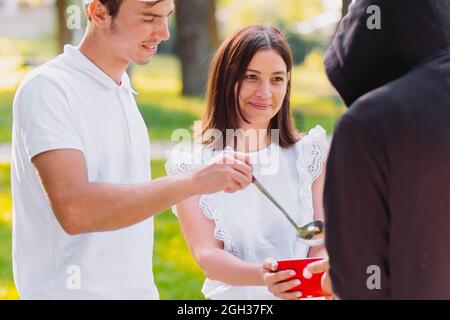 Foto von zwei freundlichen Menschen, einer jungen lächelnden Frau und einem Mann, der einem obdachlosen Mann Nahrung gab, Freiwilligenmission. Stockfoto