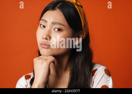 Junge nachdenkliche asiatische Frau in Sommerkleidung, die auf orangefarbenem Hintergrund steht und die Kamera anschaut Stockfoto
