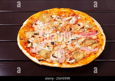 Auf einem Pappteller auf einem Holztisch wird eine ganze rustikale Pizza mit eingelegter Gurke, Speck und Spiegeleiern serviert Stockfoto