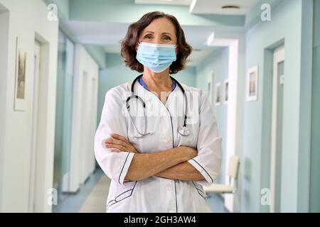 Alte Ärztin mit Gesichtsmaske und medizinischem Mantel, die in der Krankenhaushalle steht. Stockfoto