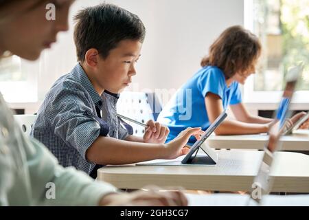 Fokussierter asiatischer Schuljunge mit digitalem Tablet in der Klasse im Klassenzimmer. Stockfoto