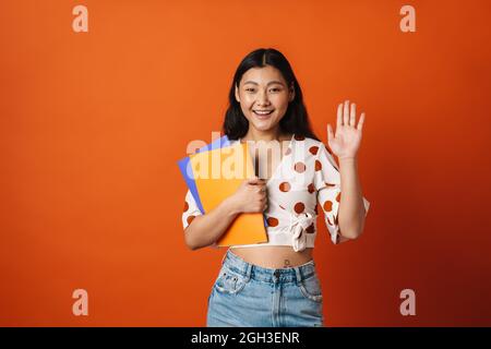 Lächelnde junge asiatische Frau Student stehend hält Lehrbücher über roten Wand Hintergrund winkende Hand Stockfoto