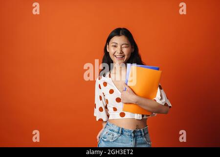 Lächelnde junge asiatische Frau Student stehend hält Lehrbücher über roten Wand Hintergrund winken Stockfoto