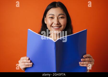 Glücklich junge Brünette asiatische Frau hält Copybook stehen über orangenen Hintergrund Stockfoto