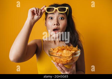 Schockiert junge asiatische Frau in 3d-Brille Film mit Schale von Potati Chips stehend über gelbem Hintergrund beobachten Stockfoto