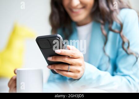 Nahaufnahme der Hände einer nicht erkennbaren Geschäftsfrau, die Kaffee trinkt, während sie auf einem Sofa sitzt, während sie ein Smartphone benutzt. Eine arbeitende Frau sendet Textnachrichten von Stockfoto